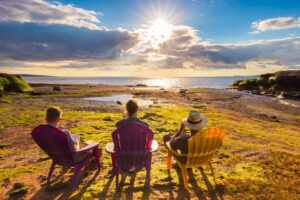 Three tourists sitting on colorful chairs enjoying a panoramic sunset view of the Atlantic Ocean tides on the green mainland and shores of Burntcoat Head Park.